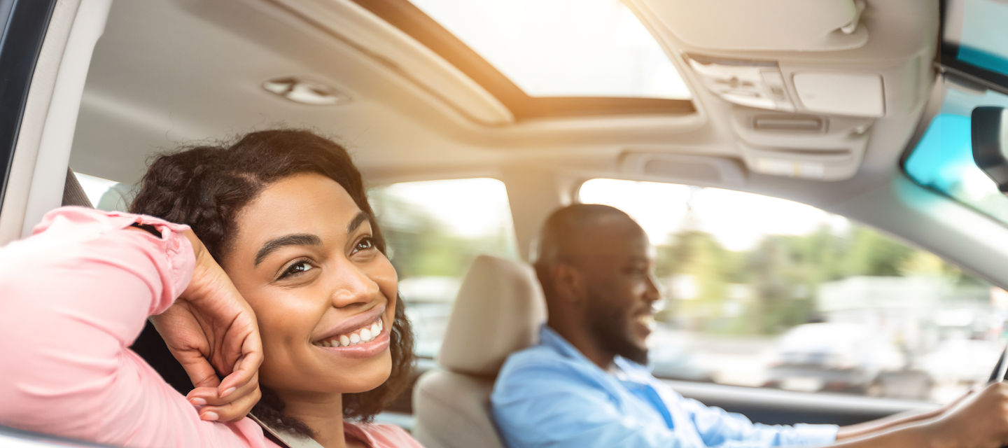 Enjoying Travel Concept. Smiling handsome African American man driving a car, selective focus on happy beautiful young woman sitting on the front passenger seat and looking out of window