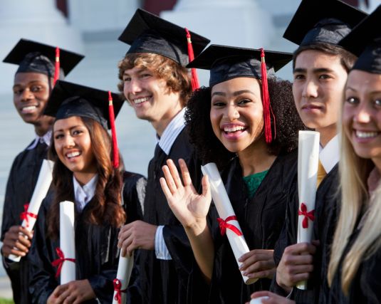 Multi-ethnic friends graduating together, in cap and gown. Main focus on African American girl in middle, waving at camera.