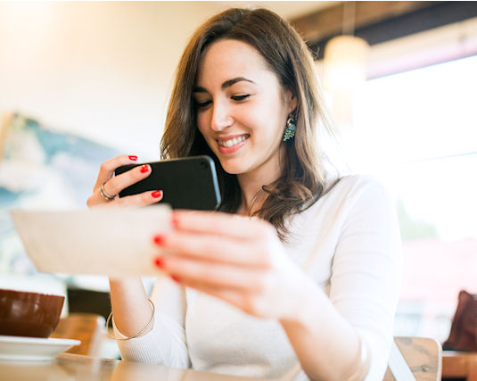 A smiling young woman takes a picture with her smart phone of a check or paycheck for digital electronic depositing, also known as 